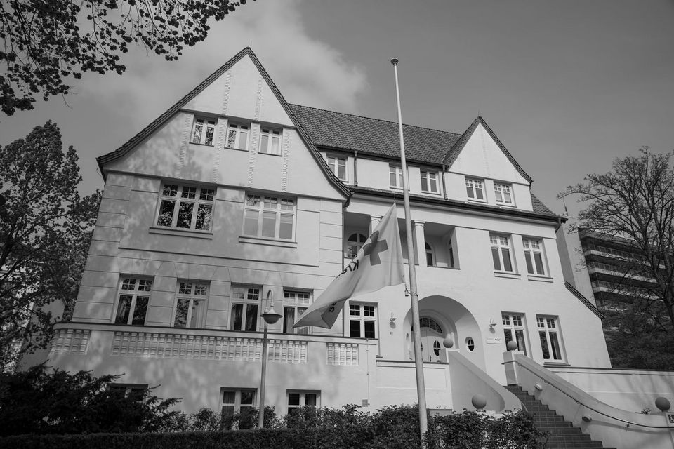 Ein schwarz-weiß-Bild zeigt die Geschäftsstelle des DRK-Landesverbandes Schleswig-Holstein. Das Gebäude ist eine große weiße Altbauvilla. Die DRK-Flagge vor dem Gebäude hängt auf Halbmast.