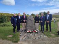 Je drei Personen stehen links und rechts eines Gedenksteins. Vor ihm liegen zwei Blumengestecke.